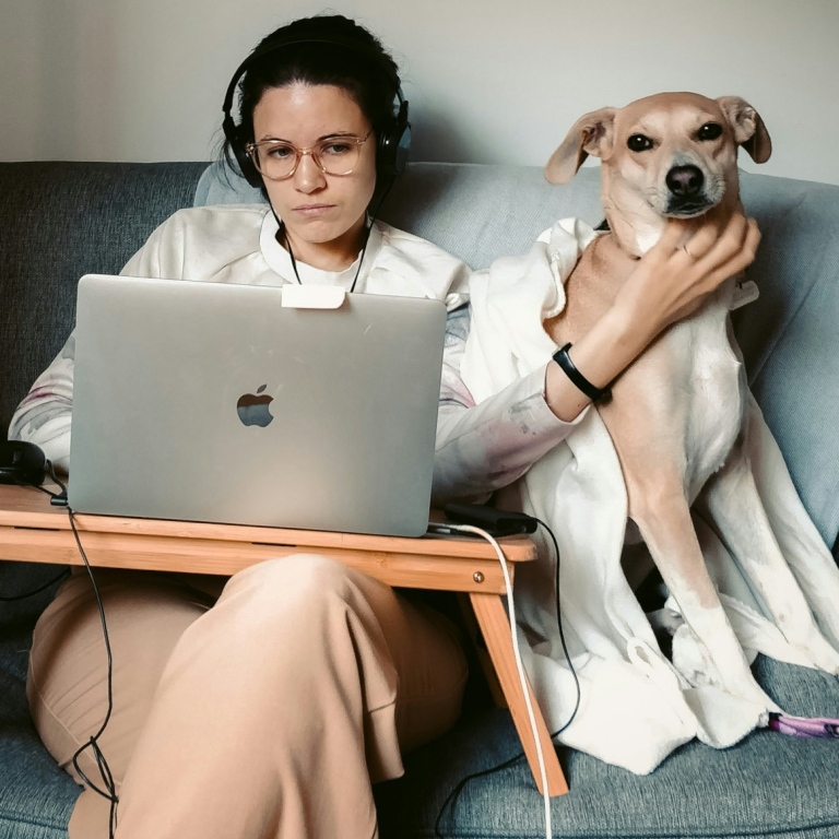 Genrebild på en kvinna med dator och hörlurar som sitter i en soffa med sin hund.