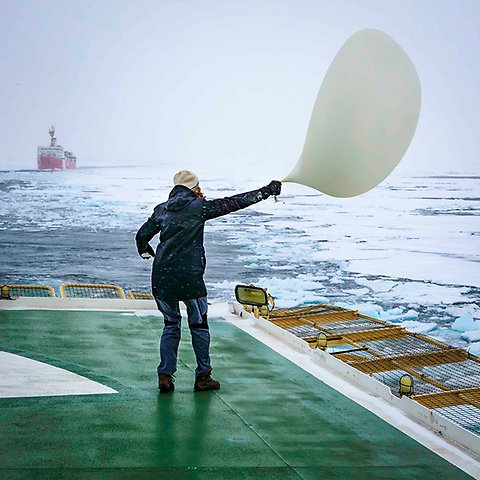 A person standing on deck on icebreaker Oden holding a weather balloon. It's windy, a bit rough sea, and another icebreaker in the far background.