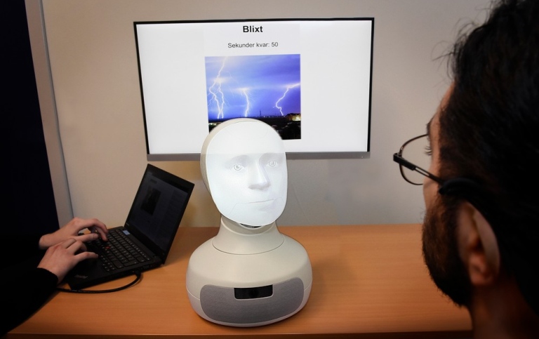En robotbyst med ett ansikte framför en datorskräm och en person som pratar med den.