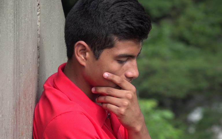 A dark-haired boy in red shirt sitting down, looking sad, with his left hand on his right cheek. 