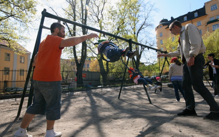 Two fathers on parental leave play with their children on the swings in a playground in Stockholm.