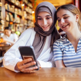 Två glada flickor sitter vid ett bord och tittar på samma mobiltelefon.