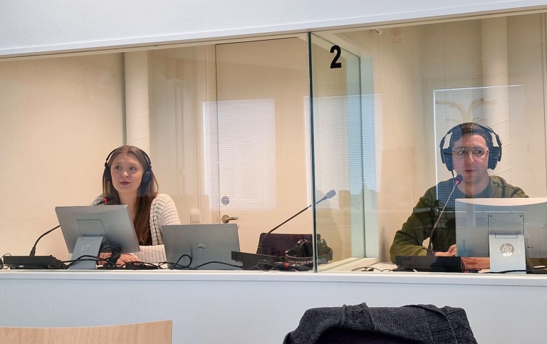 Två personer sitter i varsin tolkkabin med hörlurar på sig, mikrofonstativ och datorer, i Tolkstudion.