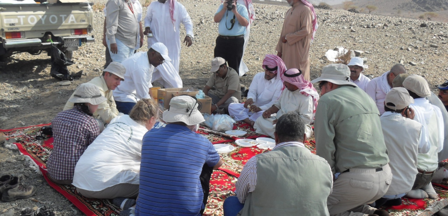 people sitting on a big matt on the deserts, eating lunch 