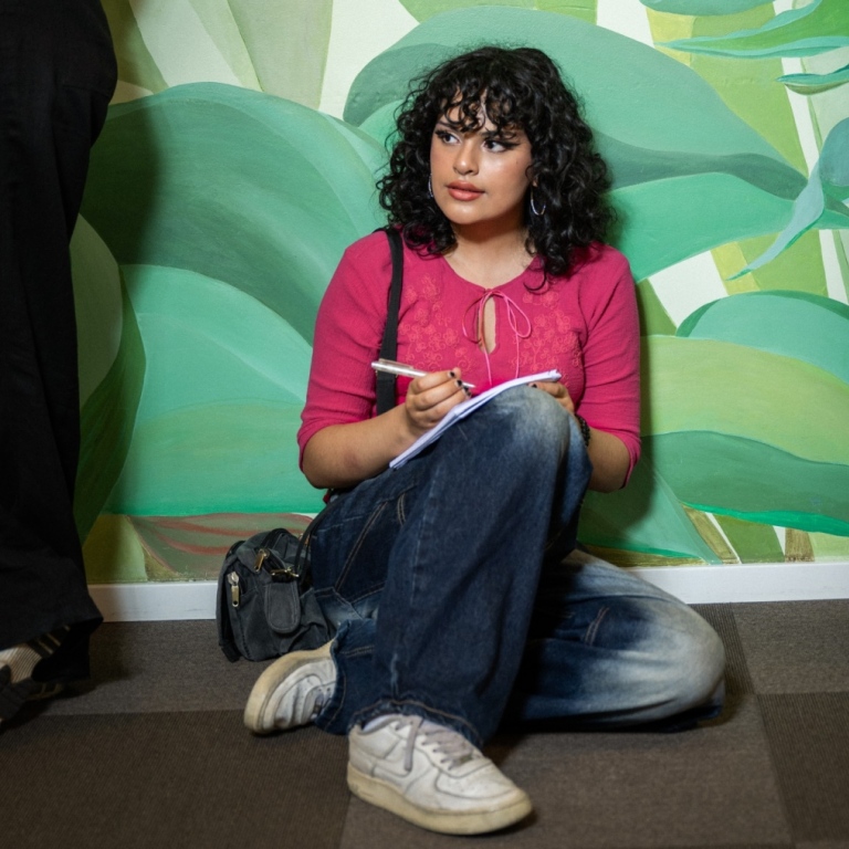 Kvinna sitter på golvet och lutar sig mot en vägg och skriver i en bok. Gröna växter målade på vägg