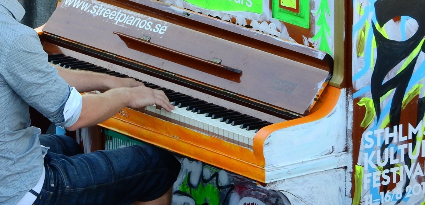 En person i grå skjorta spelar på ett piano målat i starka färger. Utomhus. 