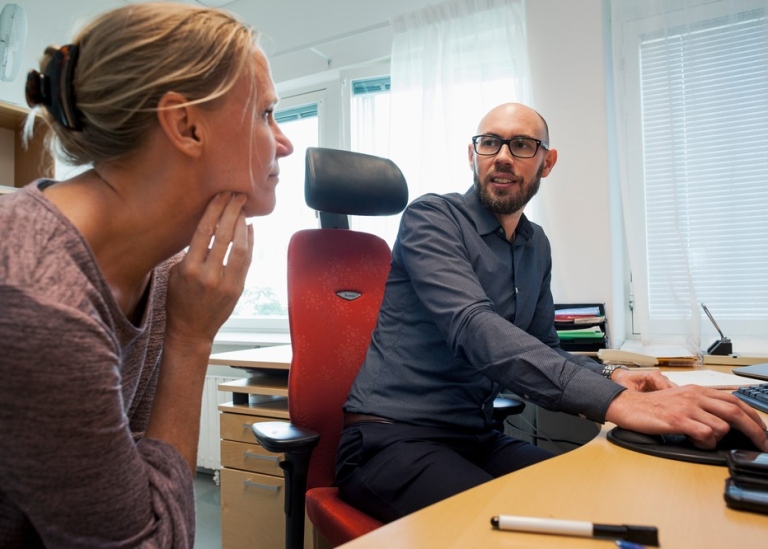 Två kollegor, kvinna och man, sitter vid skrivbord och pratar. Foto: Jens Olof Lasthein