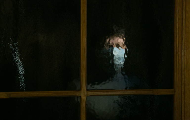 Bild på fönster och inomhus ses en person som bär munskydd titta ut
