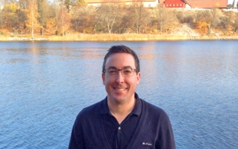 Porträttfoto av leende man med glasögon vid en sjö.