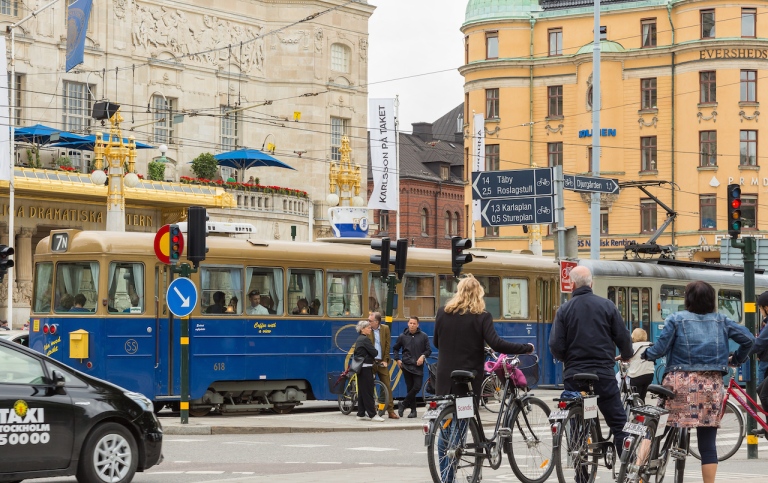 Genrebild som visar trafik i Stockholm med människor, cyklar, bilar och spårvagnar. 