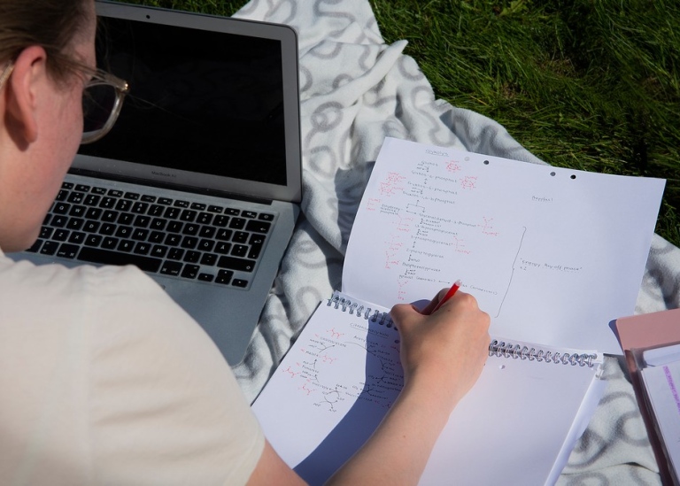 Student sitter med laptop på gräsmatta och skriver i block. Foto: Ingmarie Andersson