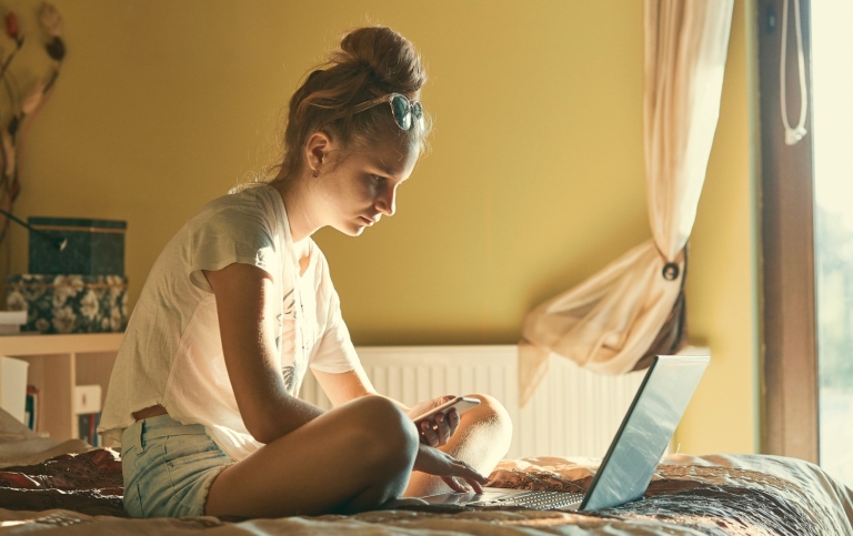 Tonåring sitter på sin säng med laptop och mobil