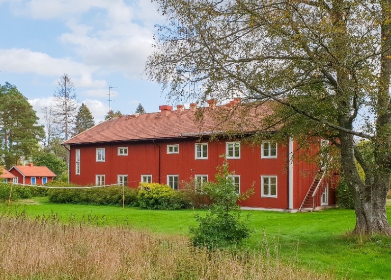 Röd tvåvåningsbyggnad med vita knutar med gräsmatta och träd i omgivningen.