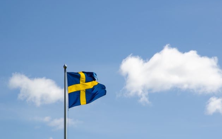 Svenska flaggan på helstång under blå himmel med enstaka stackmoln.