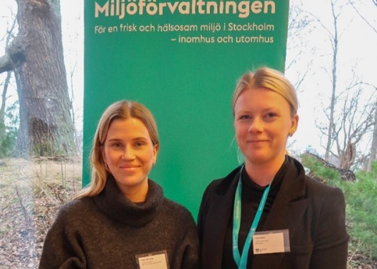 Two women with name tags stand in front of a sign with the heading "Miljöförvaltningen"
