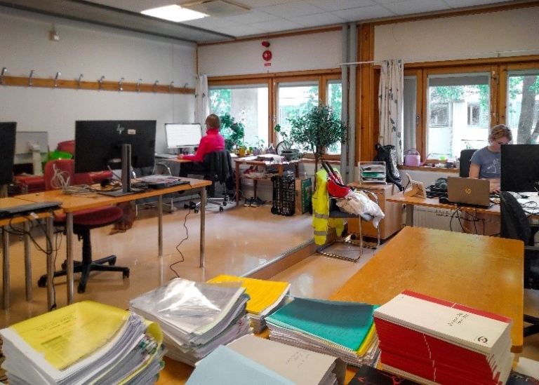 Ett stort rum fullt av skrivbord och högar med papper och saker. Två personer jobbar mitt i röran.
