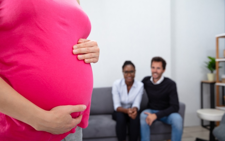 gravid kvinna i förgrunden och ett par som sitter i en soffa i bakgrunden, tittandes på den gravida 