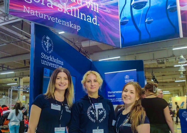 Tre kvinnor står i en mässhall under en stor skylt för Naturvetenskap vid Stockholms universitet