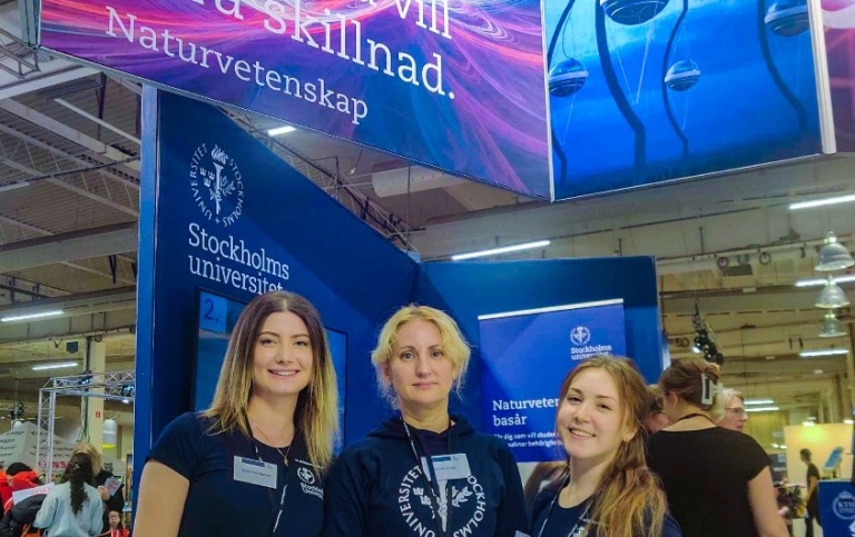 Tre kvinnor står i en mässhall under en stor skylt för Naturvetenskap vid Stockholms universitet