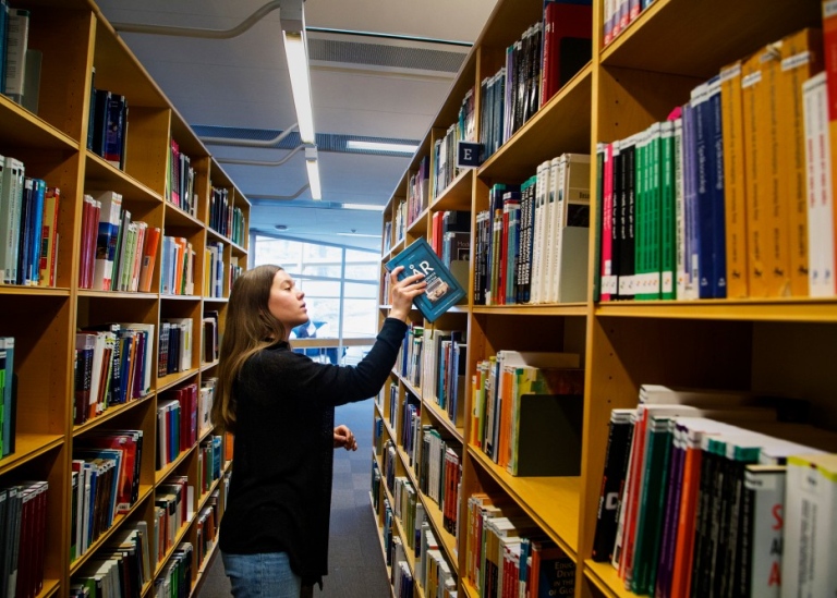 Student som håller en kursbok vid hyllorna för kurslitteratur på biblioteket