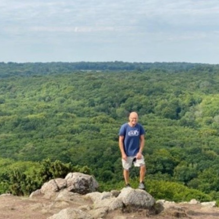 En man i tshirt och shorts står på en sten uppe på en höjd, med grönskande utsikt.