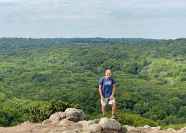 En man i tshirt och shorts står på en sten uppe på en höjd, med grönskande utsikt.