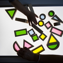 Childrens' hands playing with neon coloured geometrical puzzle pieces on a light board