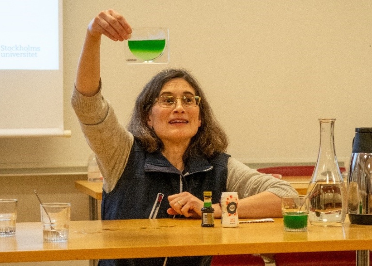 Kvinna vid ett bord med salt, matlagningsfärg och vatten, håller upp glasbehållare med grön vätska.
