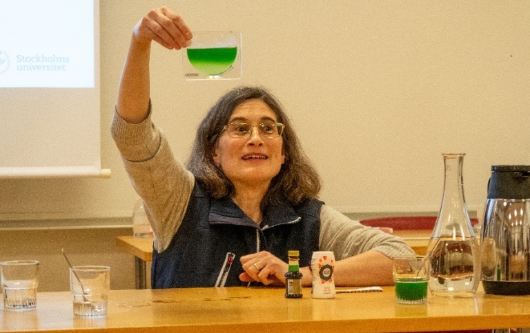 Kvinna vid ett bord med salt, matlagningsfärg och vatten, håller upp glasbehållare med grön vätska.