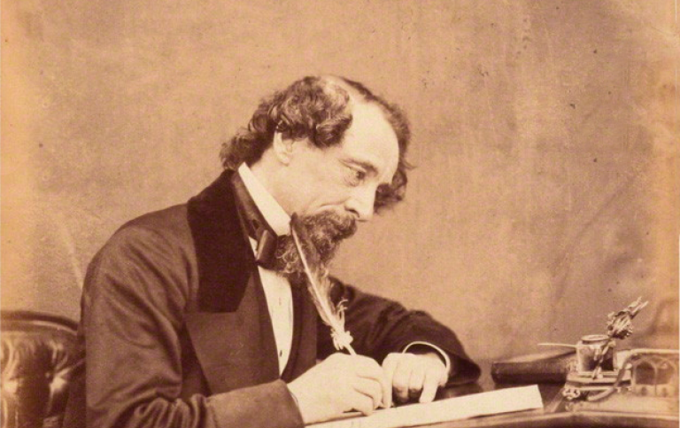 Foto föreställande Charles Dickens som sitter och skriver vid ett skrivbord