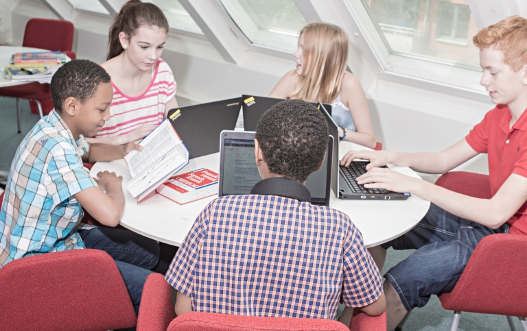 Fem elever sitter runt ett bord och arbetar med skolmaterial: böcker och datorer.