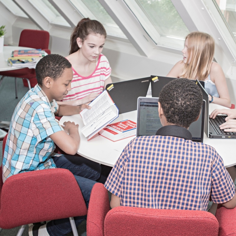 Fem elever sitter runt ett bord och arbetar med skolmaterial: böcker och datorer.