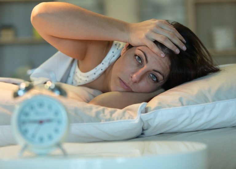 Kvinna som ligger i sängen, ser trött ut och tittar på klockan.