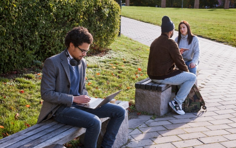 Student framför laptop medan två andra studenter pratar utomhus på campus.