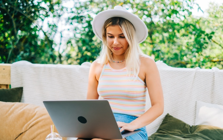 Ung sommarklädd kvinna sitter utomhus och skriver på laptop i knä. 