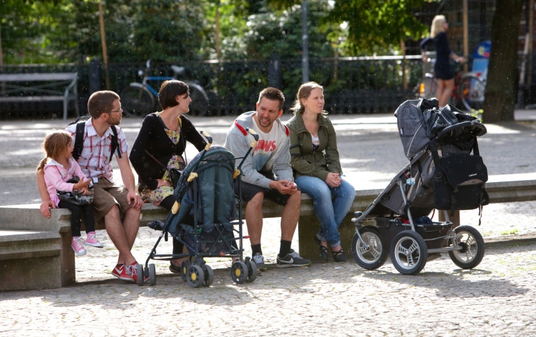 Four parents sit on a bench in a park and talk with a pram in front of them.