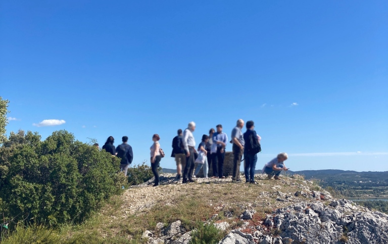 En grupp människor står på ett berg med utsikt över en dal. Blå himmer och sol.