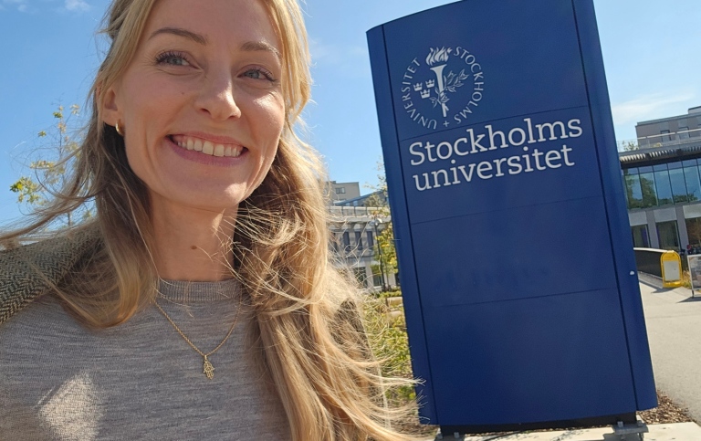 Sofia Lindblom vid blå skylt med Stockholms universitets logotyp framför Campus Albano.