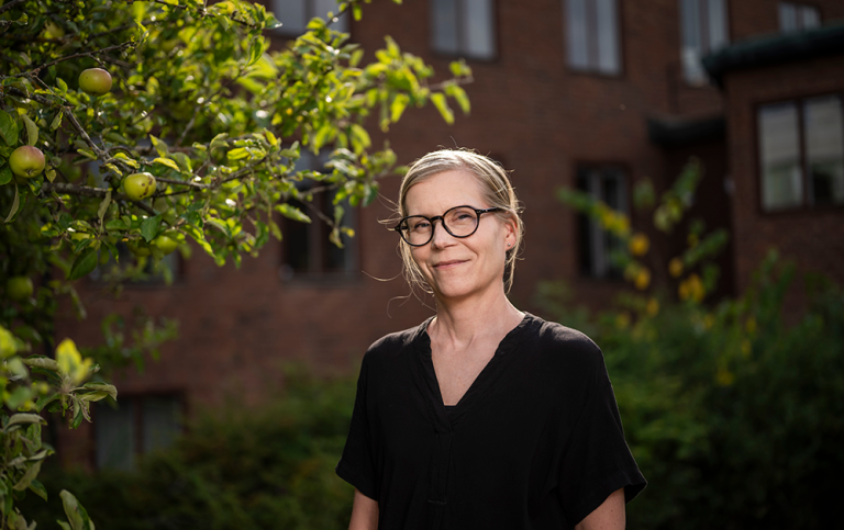 Annika Berg. Foto: Johanna Säll / Stockholms universitet Annika Berg står bredvid ett äppelträd. I bakgrunden syns en brun tegelbyggnad i tre våningar